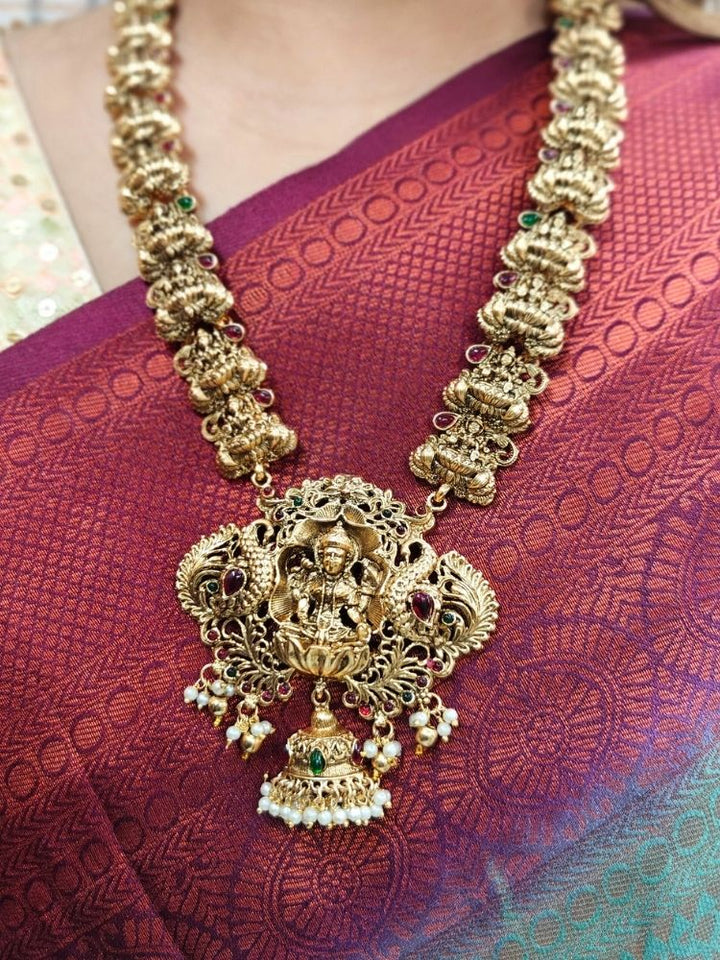 A woman in a maroon saree with gold embroidery and a cream blouse, adorned with traditional South-Indian jewelry, including the Vibhuti Temple Necklace Set featuring an ornate Laxmi necklace and matching temple jhumka earrings.
