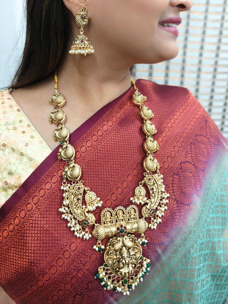 A woman wearing a maroon and teal silk saree, gold earrings, and the Vidya Temple Necklace Set—a grand gold necklace with intricate designs and pearl accents—perfect for a wedding ceremony.  Perfect necklace to add to your temple necklace collection. 