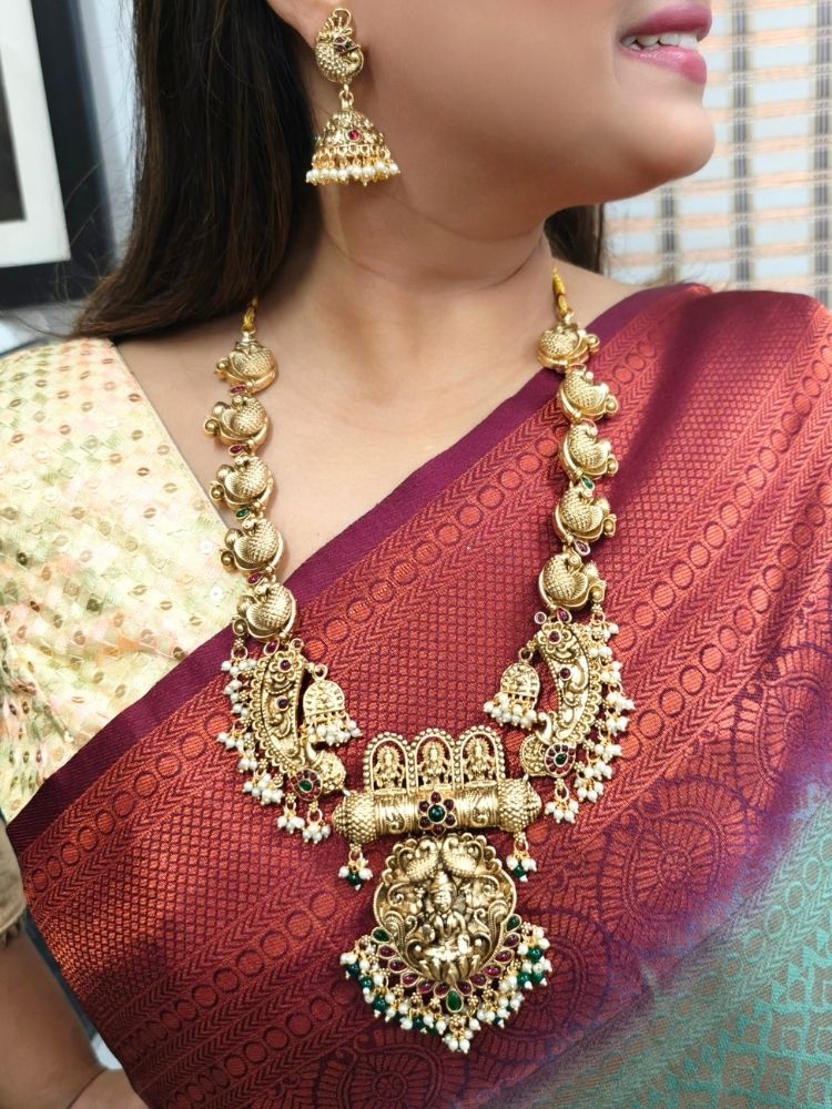 A woman wearing a maroon and teal silk saree, gold earrings, and the Vidya Temple Necklace Set—a grand gold necklace with intricate designs and pearl accents—perfect for a wedding ceremony.  Perfect necklace to add to your temple necklace collection. 