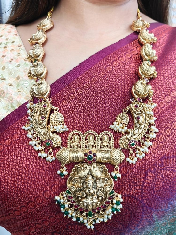 A woman wearing a maroon and teal silk saree, gold earrings, and the Vidya Temple Necklace Set—a grand gold necklace with intricate designs and pearl accents—perfect for a wedding ceremony.  Perfect necklace to add to your temple necklace collection. 