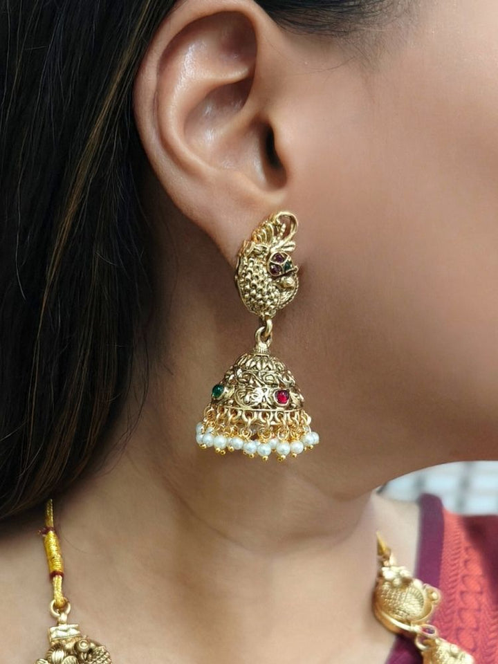 A woman wearing a maroon and teal silk saree, gold earrings, and the Vidya Temple Necklace Set—a grand gold necklace with intricate designs and pearl accents—perfect for a wedding ceremony.  Perfect necklace to add to your temple necklace collection. 