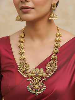 A woman wearing the Vidya Temple Necklace Set featuring a traditional gold Long necklace and matching earrings with Goddess Laxmi in the middle, along with pink and green colored stones, Pearls and Mehendi Polish. Wedding guest look completed with a temple necklace set. Close-up of detailed temple carvings in artificial wedding jewelry.