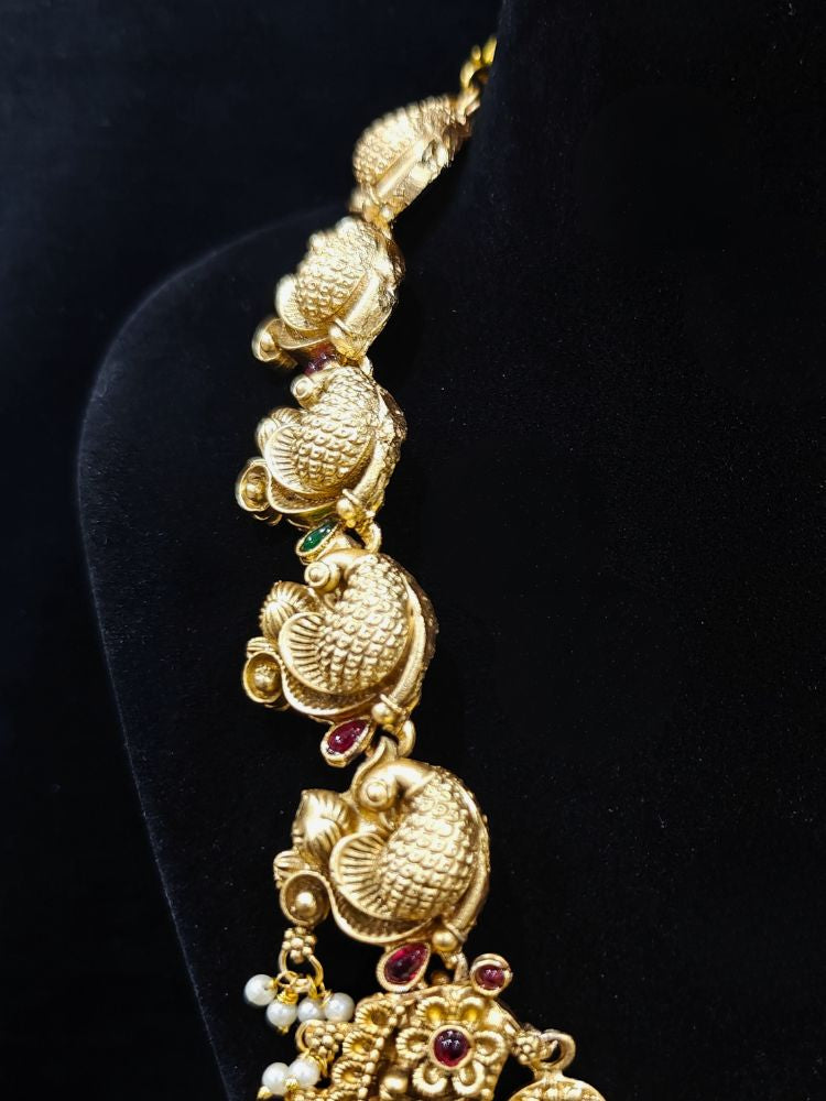 The Vidya Temple Necklace Set featuring a traditional gold Long necklace and matching earrings with Goddess Laxmi in the middle, along with pink and green colored stones, Pearls and Mehendi Polish. Wedding guest look completed with a temple necklace set. Close-up of detailed temple carvings in artificial wedding jewelry.