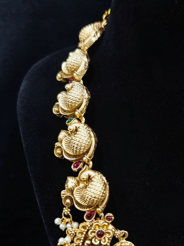 The Vidya Temple Necklace Set featuring a traditional gold Long necklace and matching earrings with Goddess Laxmi in the middle, along with pink and green colored stones, Pearls and Mehendi Polish. Wedding guest look completed with a temple necklace set. Close-up of detailed temple carvings in artificial wedding jewelry.
