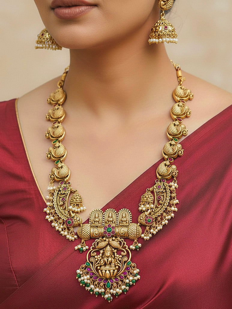 A woman wearing the Vidya Temple Necklace Set featuring a traditional gold Long necklace and matching earrings with Goddess Laxmi in the middle, along with pink and green colored stones, Pearls and Mehendi Polish. Wedding guest look completed with a temple necklace set. Close-up of detailed temple carvings in artificial wedding jewelry.