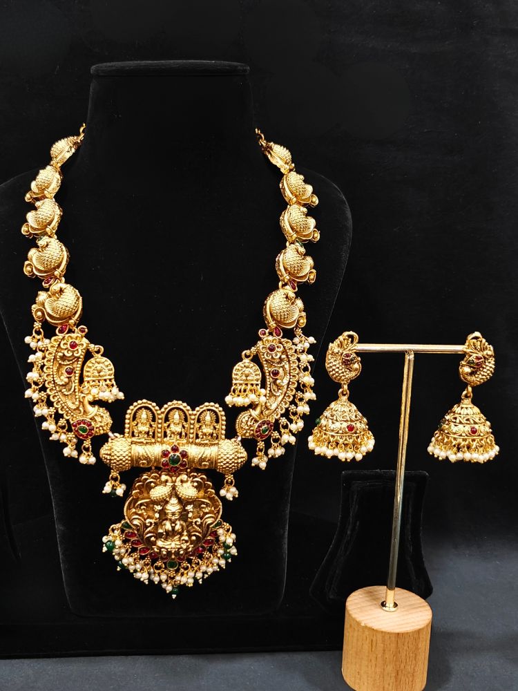 The Vidya Temple Necklace Set featuring a traditional gold Long necklace and matching earrings with Goddess Laxmi in the middle, along with pink and green colored stones, Pearls and Mehendi Polish. Wedding guest look completed with a temple necklace set. Close-up of detailed temple carvings in artificial wedding jewelry.