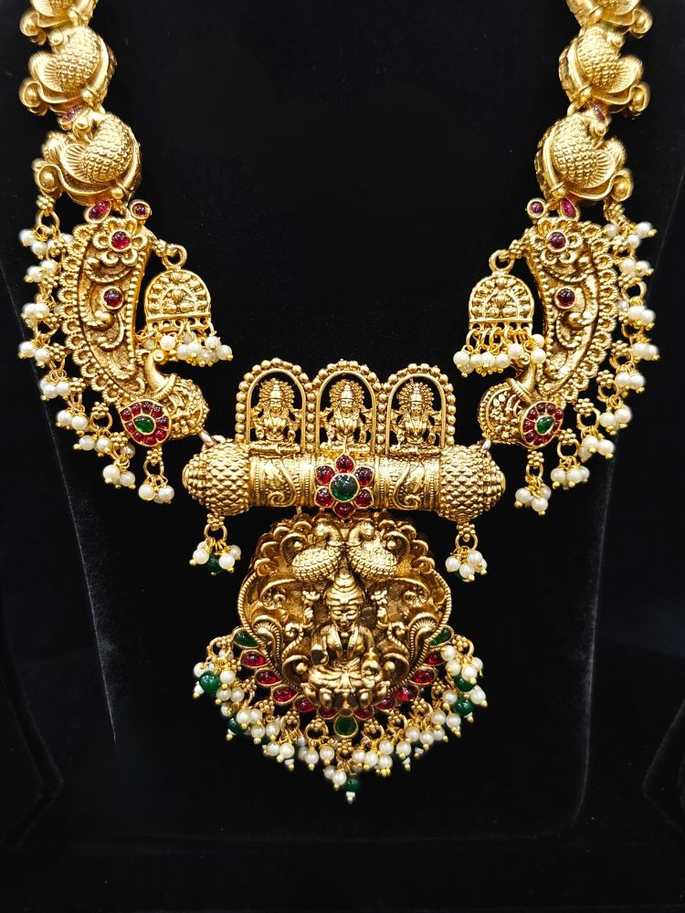 The Vidya Temple Necklace Set featuring a traditional gold Long necklace and matching earrings with Goddess Laxmi in the middle, along with pink and green colored stones, Pearls and Mehendi Polish. Wedding guest look completed with a temple necklace set. Close-up of detailed temple carvings in artificial wedding jewelry.