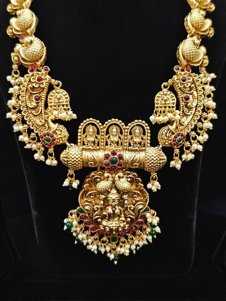 The Vidya Temple Necklace Set featuring a traditional gold Long necklace and matching earrings with Goddess Laxmi in the middle, along with pink and green colored stones, Pearls and Mehendi Polish. Wedding guest look completed with a temple necklace set. Close-up of detailed temple carvings in artificial wedding jewelry.