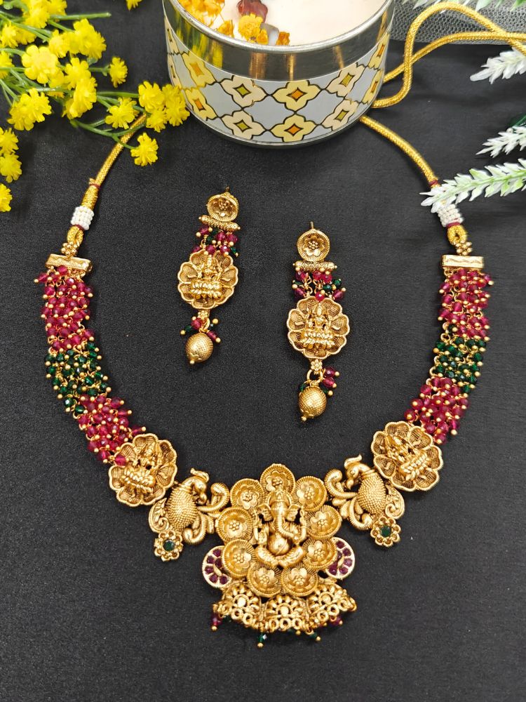 The Vinayaka Necklace Set features a gold temple necklace and ethnic earrings with intricate designs, red and green beads, floral motifs, and a central Lord Ganesha pendant, displayed on black with yellow flowers and a decorative bowl.