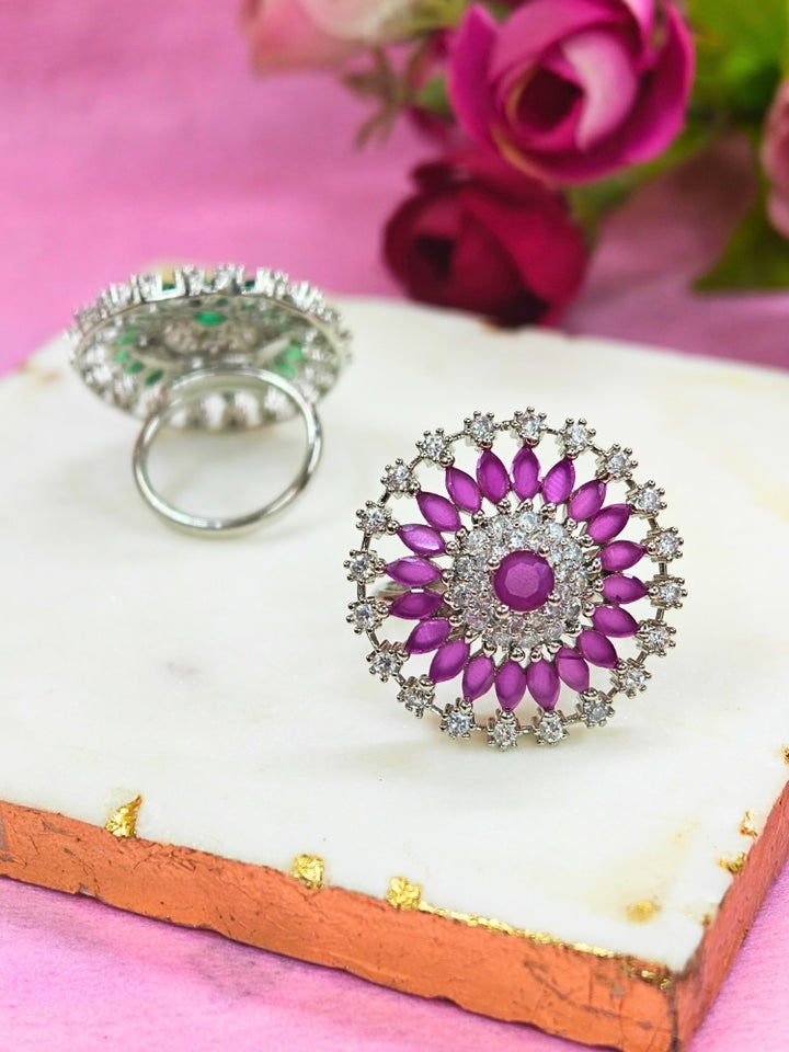 The Wallace Pink Ring features a circular design with a central purple gemstone, surrounded by American diamonds and marquise-shaped purple stones, set against a pink and white background adorned with roses. Perfect American Diamond cocktail ring. 