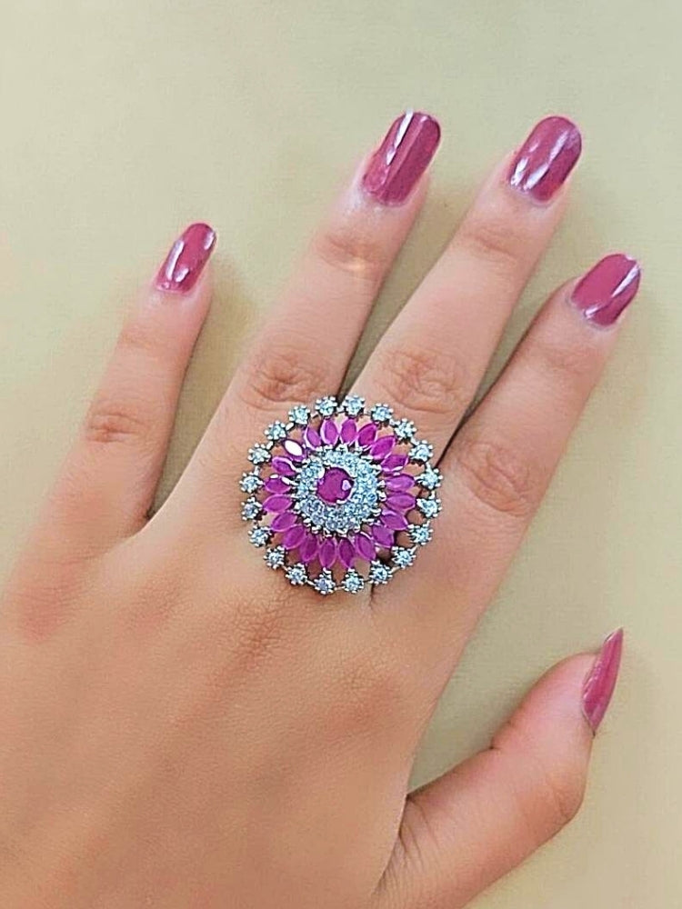 The Wallace Pink Ring features a circular design with a central purple gemstone, surrounded by American diamonds and marquise-shaped purple stones, set against a pink and white background adorned with roses. Perfect American Diamond cocktail ring. 