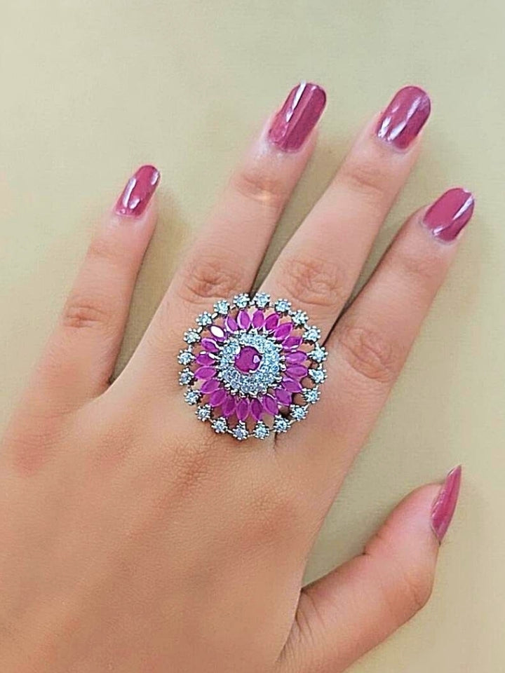 The Wallace Pink Ring features a circular design with a central purple gemstone, surrounded by American diamonds and marquise-shaped purple stones, set against a pink and white background adorned with roses. Perfect American Diamond cocktail ring. 