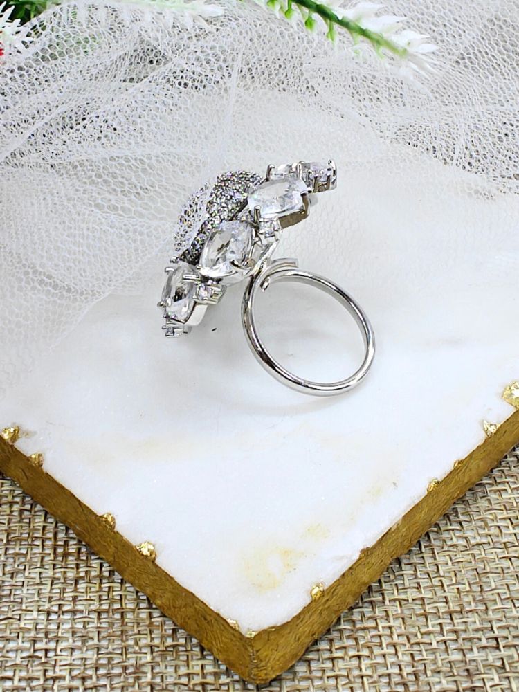The White Aster Ring is a round silver piece with a sparkling, round-shaped center made with white American diamonds, surrounded by petal shaped White gemstones, along with an adjustable band. It’s shown on a textured surface with white netting and green leaves in the background.