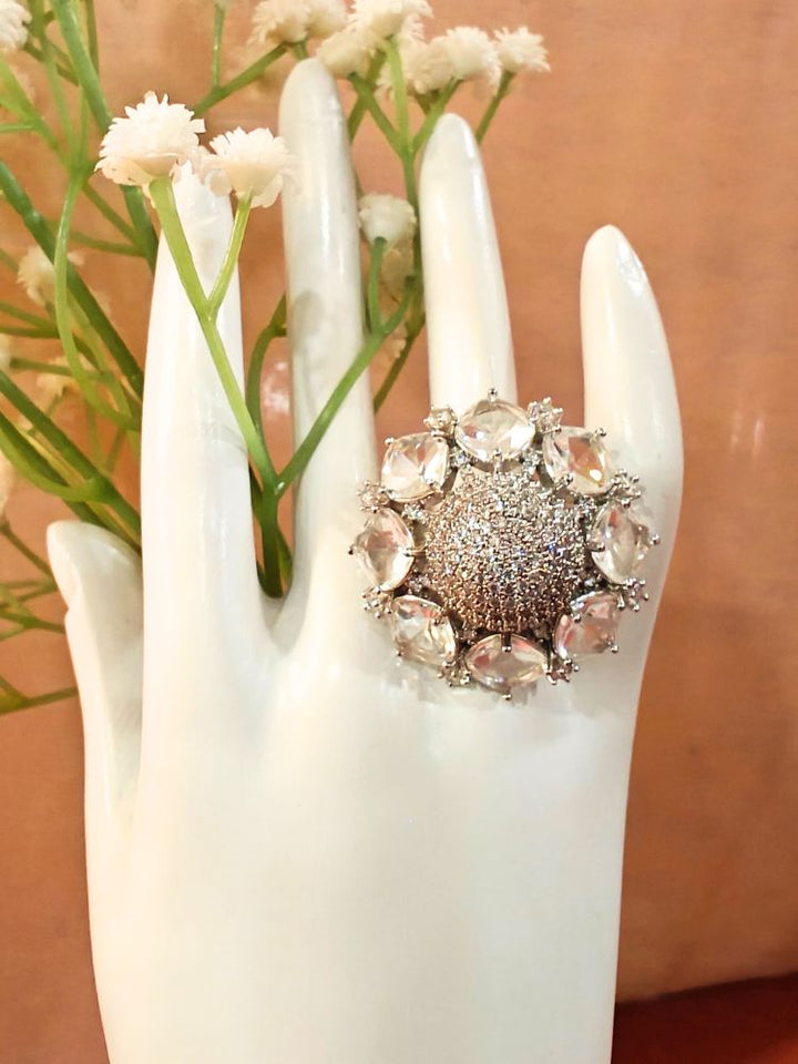 The White Aster Ring is a round silver piece with a sparkling, round-shaped center made with white American diamonds, surrounded by petal shaped White gemstones, along with an adjustable band. It’s shown on a White Hand Mannequin.