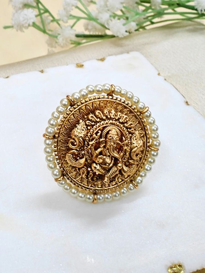 The White Bheema Ring is a round, gold-toned adjustable ring featuring Ganeshji made in intricate detailing at the center, encircled by Pearls. Displayed on white cloth with small flowers, its ideal for enhancing your wedding ceremony outfit.