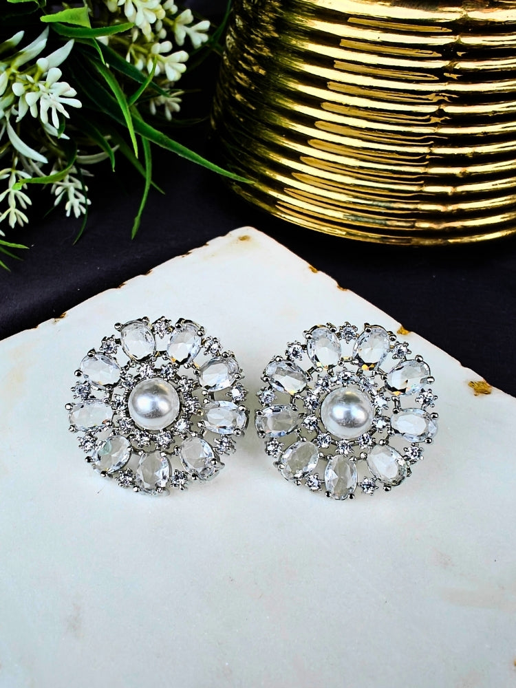 The White Blossom Earrings showcase a central white pearl encircled by alternating White stones and American diamonds, elegantly displayed on White Marble with green leaves and a Golden Accent.