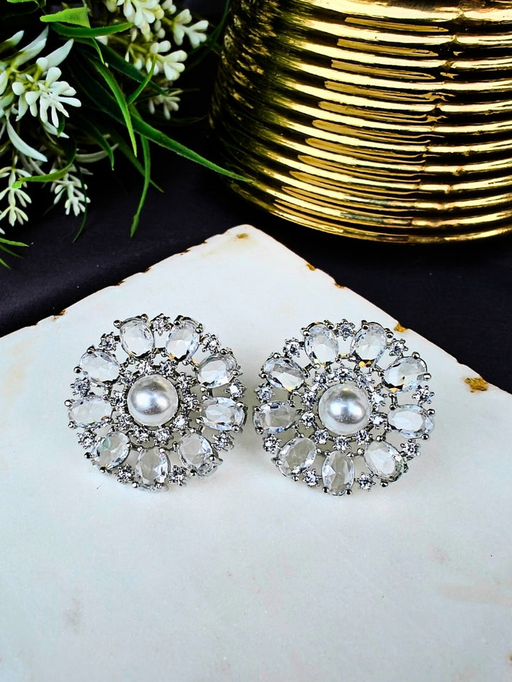 The White Blossom Earrings showcase a central white pearl encircled by alternating White stones and American diamonds, elegantly displayed on White Marble with green leaves and a Golden Accent.