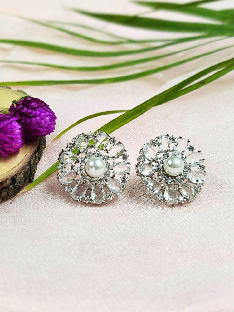 The White Blossom Earrings showcase a central white pearl encircled by alternating White stones and American diamonds, elegantly displayed on soft pink fabric with green leaves and a purple flower.
