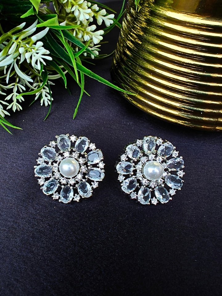 The White Blossom Earrings showcase a central white pearl encircled by alternating White stones and American diamonds, elegantly displayed on Black fabric with green leaves and a Golden Accent.