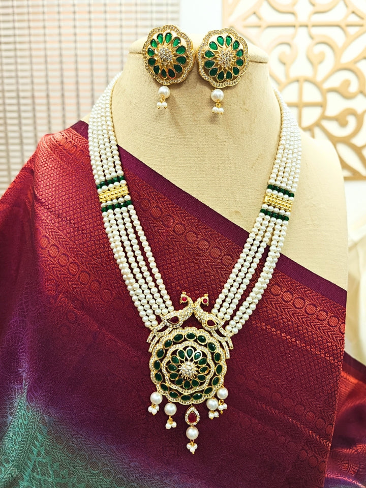 A mannequin showcases the White Gayatri Necklace Set: a  White multi strand necklace made with Pearls, along with an American Diamond pendant with peacock design with pearls hanging at the bottom. The necklace comes with matching earrings, styled with a Maroon saree—ideal for wedding ceremonies.
