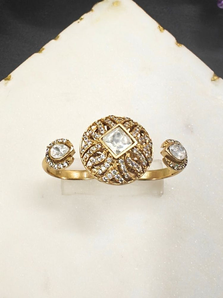 The White Octavia Ring features a gold band with a round, ornate centerpiece set with a clear American Diamond and two smaller White gemstones on the side, displayed on a white surface with gold trim. Multi-finger ring, great for cocktail parties. 