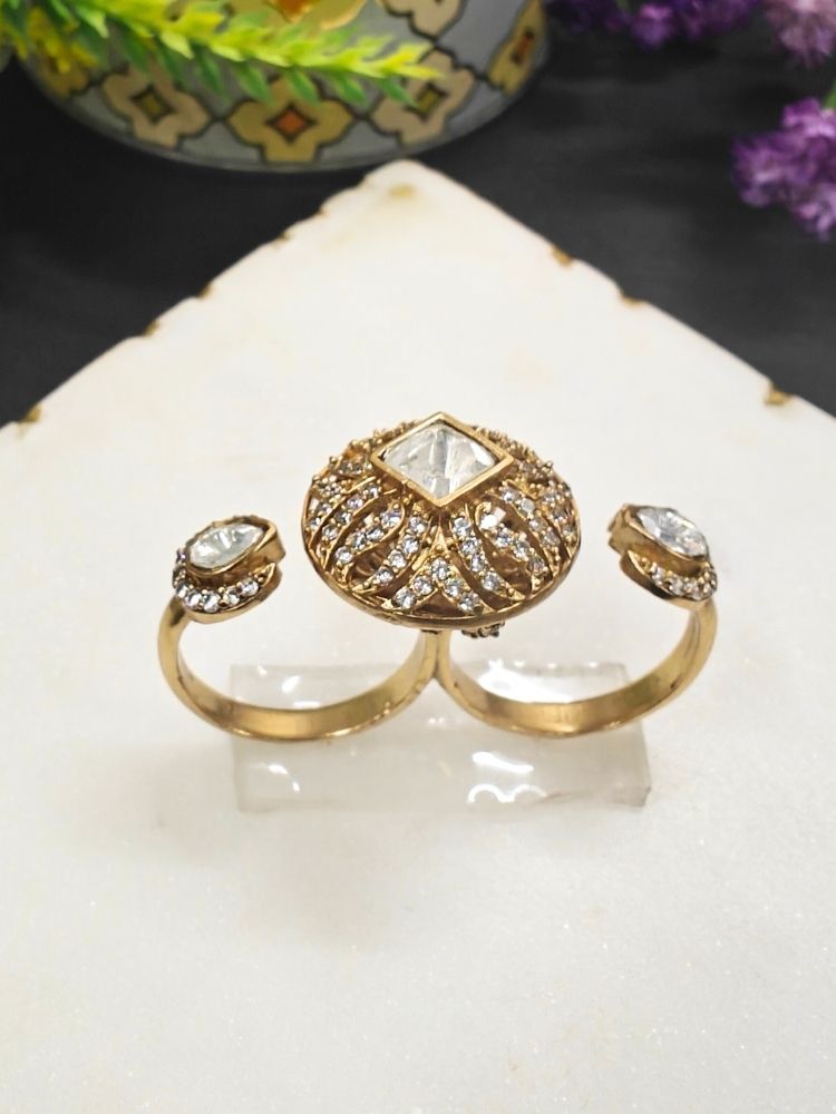 The White Octavia Ring features a gold band with a round, ornate centerpiece set with a clear American Diamond and two smaller White gemstones on the side, displayed on a white surface with gold trim. Multi-finger ring, great for cocktail parties. 