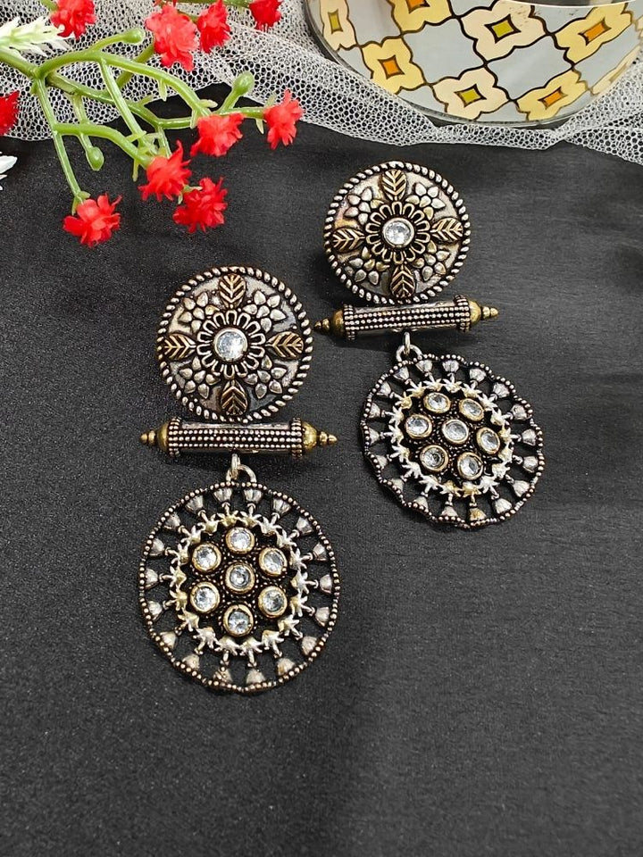 The White Sheetal Earrings feature intricate circular brass designs with embedded American diamonds, offering an antique oxidized look. Displayed with red flowers and a patterned container, they’re perfect for a wedding ceremony. Perfect to wear on navratri. 