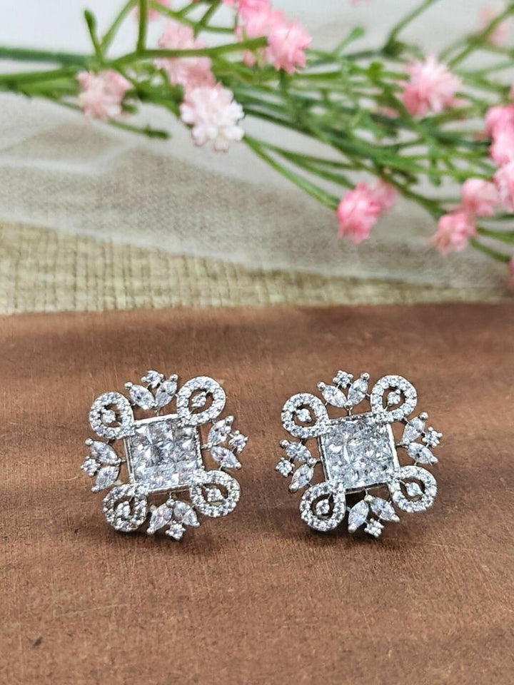 The White Zuri Earrings feature ornate White square gemstones at the center, accented by intricate silver designs and sparkling American diamonds, displayed on a brown surface with pink flowers in the background.