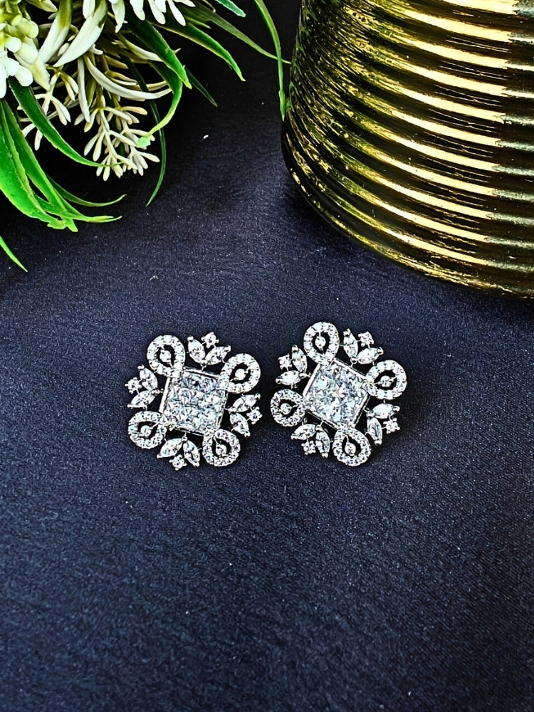 The White Zuri Earrings feature ornate White square gemstones at the center, accented by intricate silver designs and sparkling American diamonds, displayed on a Black surface with White flowers in the background.