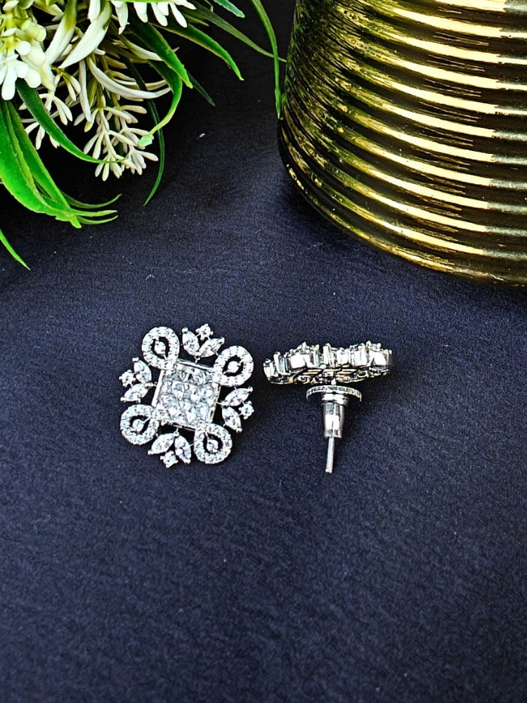 The White Zuri Earrings feature ornate White square gemstones at the center, accented by intricate silver designs and sparkling American diamonds, displayed on a Black surface with White flowers in the background.