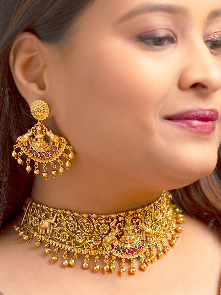 A woman wearing the Yagnika Choker Necklace Set featuring a traditional gold choker necklace and matching chandbali earrings with Goddess Laxmi in the middle, along with pink and green colored stones, golden elephants at the corners, paired with a pink and gold blouse—ideal party jewelry for any festive occasion.