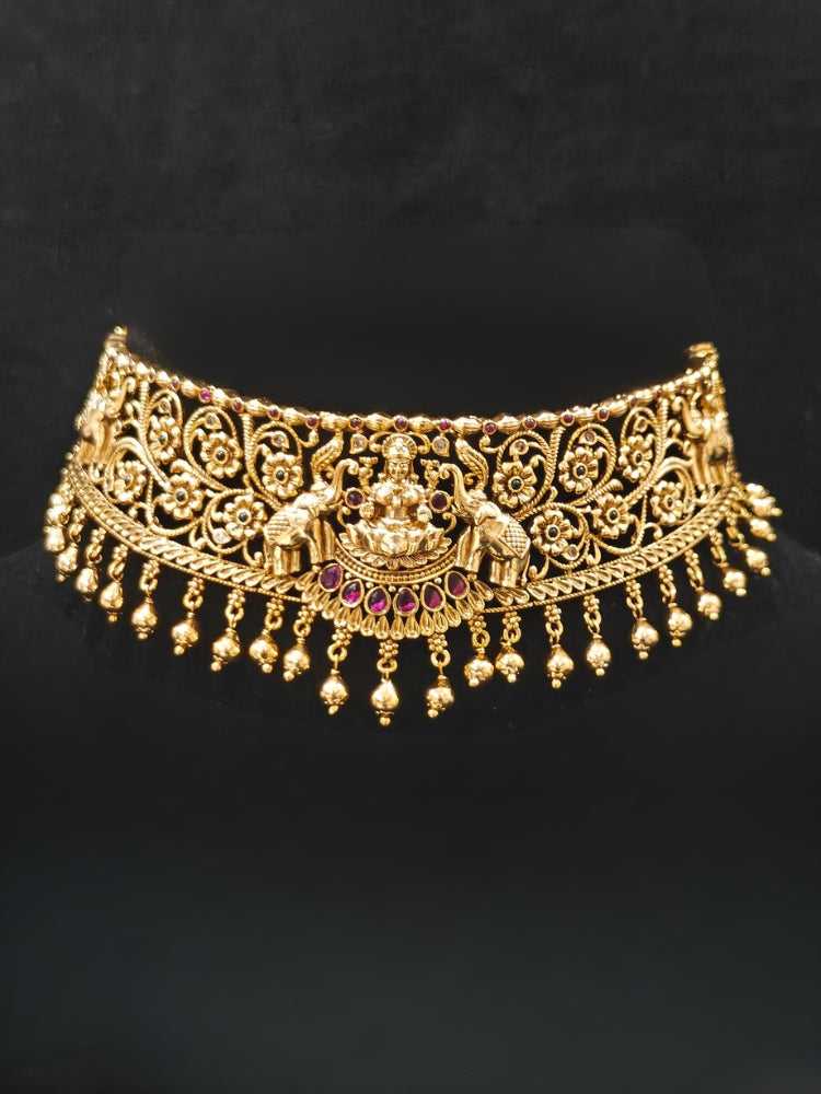 The Yagnika Choker Necklace Set featuring a traditional gold choker necklace and matching chandbali earrings with Goddess Laxmi in the middle, along with pink and green colored stones, golden elephants at the corners, paired with a pink and gold blouse—ideal party jewelry for any festive occasion.