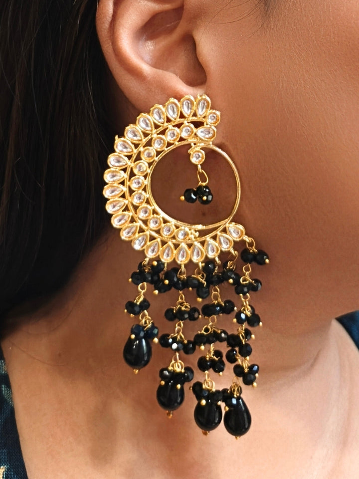 A close-up of a person wearing Yashika Black Long Earrings— large, ornate gold earrings with a  circular crescent design, made with kundan stones, along with teardrop shaped Black stones for a stunning ethnic look. Great long earrings for wedding functions
