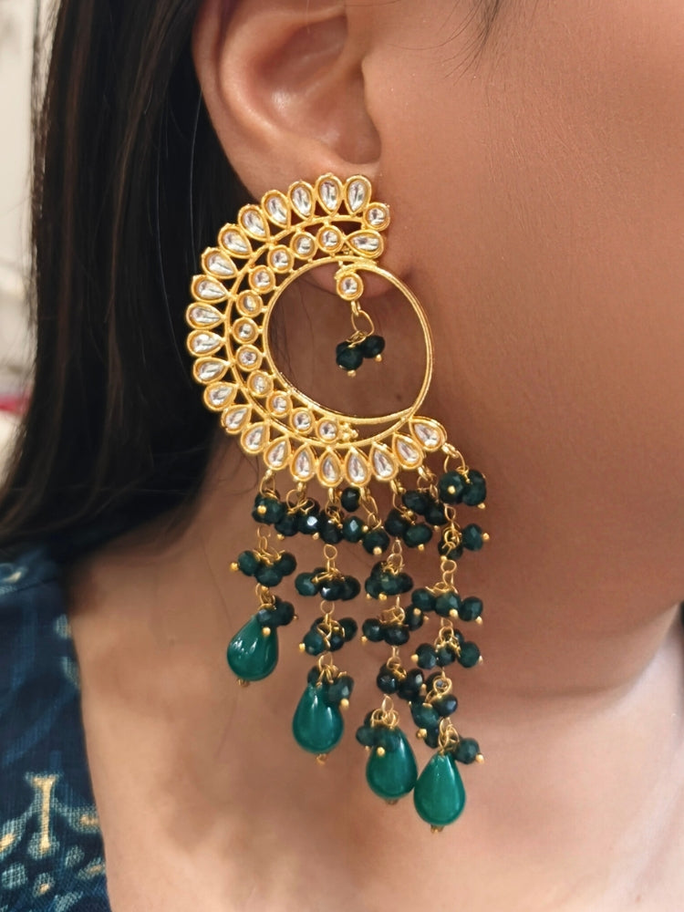 A close-up of a person wearing Yashika Green Long Earrings— large, ornate gold earrings with a  circular crescent design, made with kundan stones, along with teardrop shaped green stones for a stunning ethnic look. Great long earrings for wedding functions