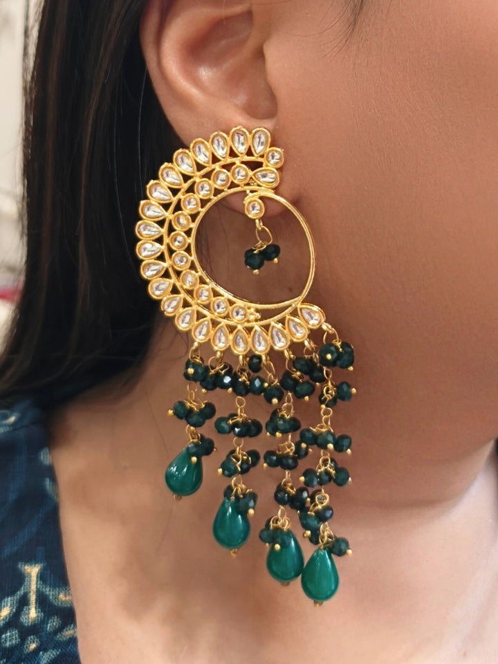 A close-up of a person wearing Yashika Green Long Earrings— large, ornate gold earrings with a  circular crescent design, made with kundan stones, along with teardrop shaped green stones for a stunning ethnic look. Great long earrings for wedding functions