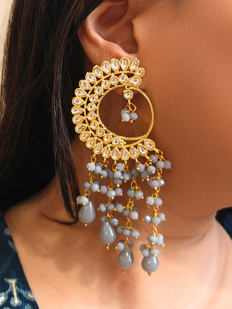 A close-up of a person wearing Yashika Grey Long Earrings— large, ornate gold earrings with a  circular crescent design, made with kundan stones, along with teardrop shaped grey stones for a stunning ethnic look. Great long earrings for wedding functions