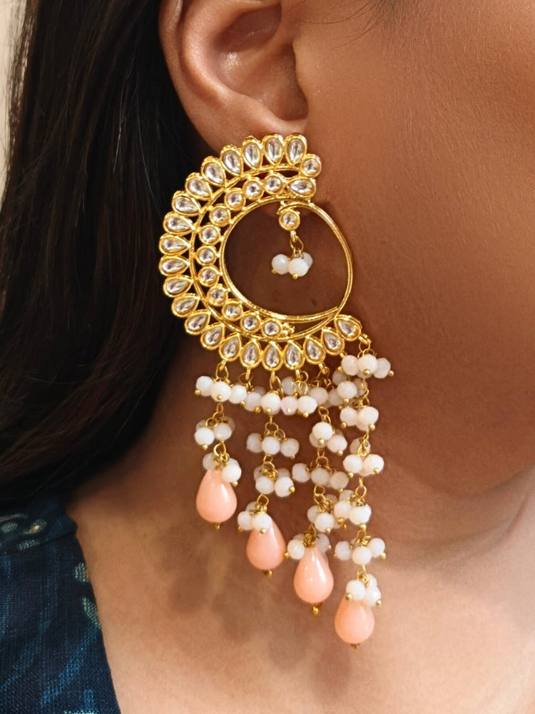 A close-up of a person wearing Yashika Peach Long Earrings— large, ornate gold earrings with a  circular crescent design, made with kundan stones, along with teardrop shaped Peach stones for a stunning ethnic look. Great long earrings for wedding functions