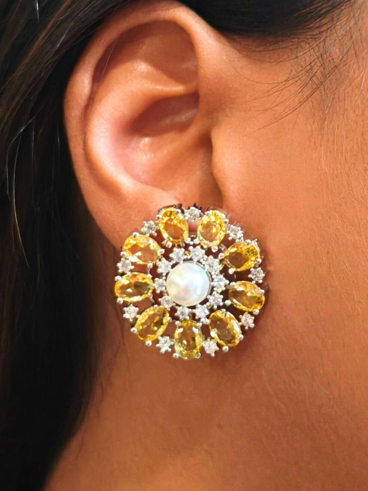 A close-up of a womans ear wearing the Yellow Blossom stud Earrings—large, circular Yellow earrings with a pearl at the center, colorful accents, and a border of sparkling American Diamonds. The stud earrings give a flower earrings feel. 