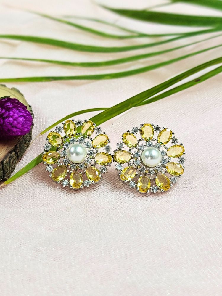 The Yellow Blossom Earrings showcase a central white pearl encircled by alternating Yellow stones and American diamonds, elegantly displayed on soft pink fabric with green leaves and a purple flower.