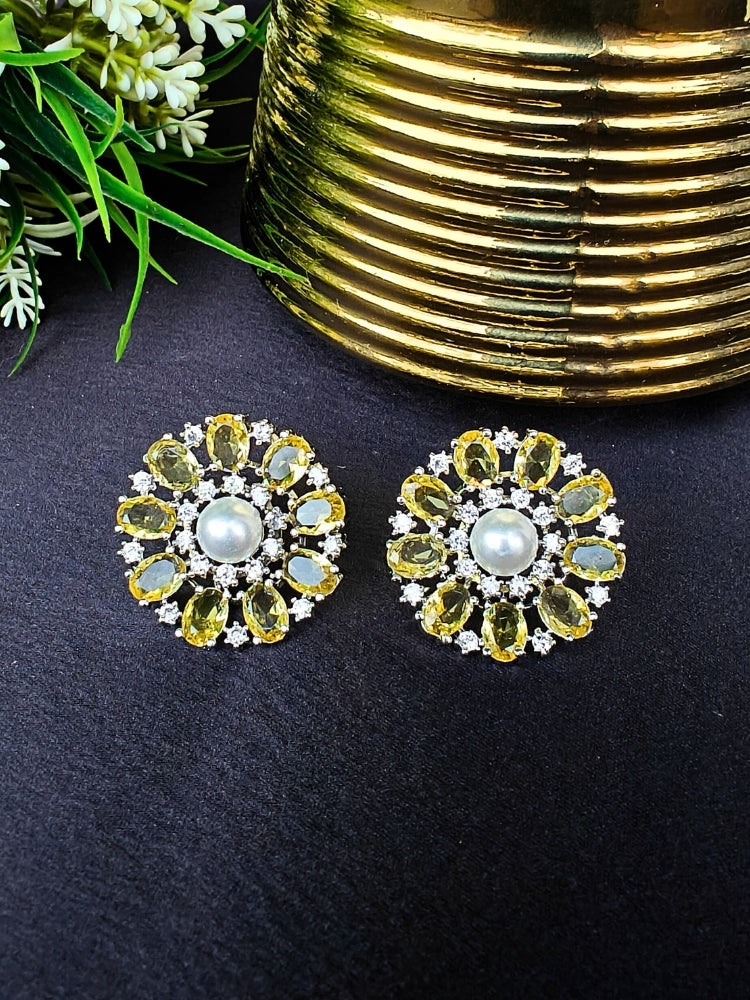 The Yellow Blossom Earrings showcase a central white pearl encircled by alternating Yellow stones and American diamonds, elegantly displayed on Black fabric with green leaves and a Golden Accent.