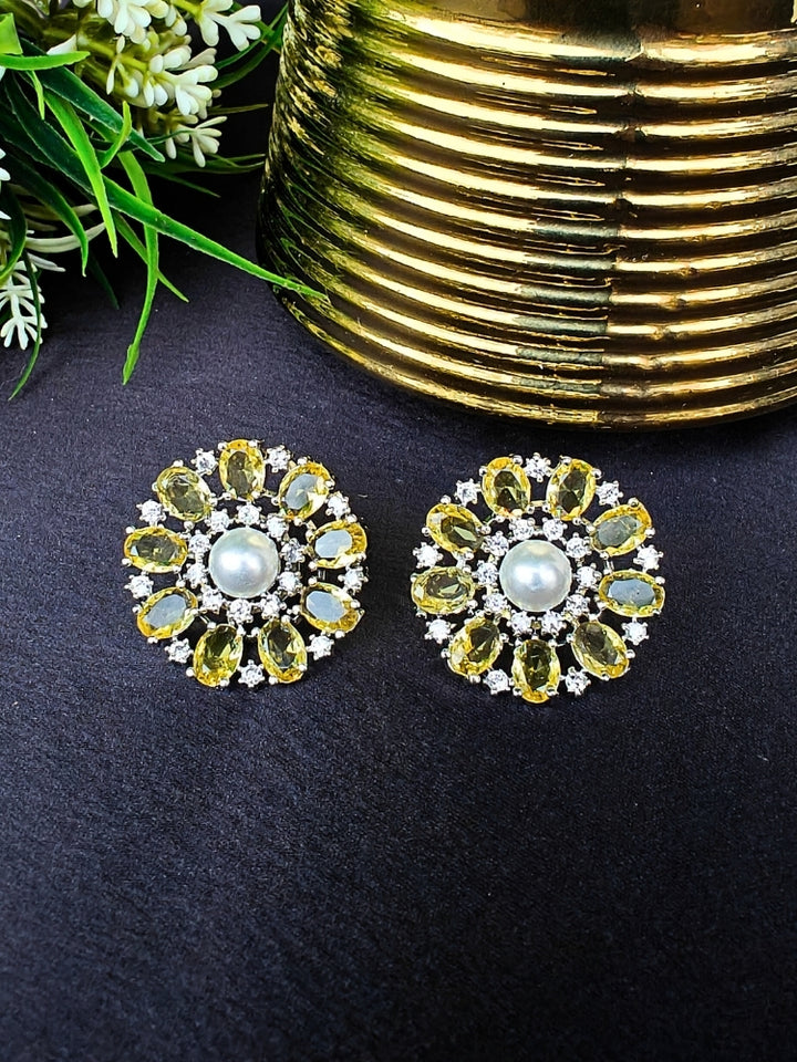 The Yellow Blossom Earrings showcase a central white pearl encircled by alternating Yellow stones and American diamonds, elegantly displayed on Black fabric with green leaves and a Golden Accent.
