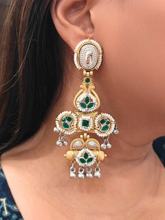 A close-up of a person wearing Zaynika Green Earrings—large, ornate brass earrings with intricate detailing, white and Green stones, and hanging silver beads. The person has long dark hair and is in a dark blue patterned top.  Long Oxidized earrings, german silver earrings, navratri earrings.  Cross shaped earrings. 