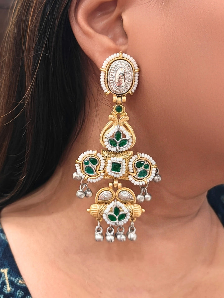 A close-up of a person wearing Zaynika Green Earrings—large, ornate brass earrings with intricate detailing, white and Green stones, and hanging silver beads. The person has long dark hair and is in a dark blue patterned top.  Long Oxidized earrings, german silver earrings, navratri earrings.  Cross shaped earrings. 