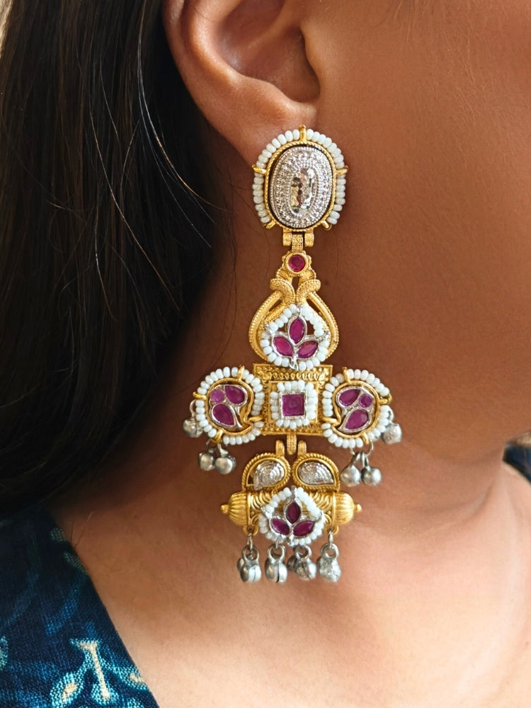 A close-up of a person wearing Zaynika Pink Earrings—large, ornate brass earrings with intricate detailing, white and pink stones, and hanging silver beads. The person has long dark hair and is in a dark blue patterned top.  Long Oxidized earrings, german silver earrings, navratri earrings.  Cross shaped earrings. 