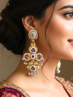 A close-up of a person wearing Zaynika Pink Earrings—large, ornate brass earrings with intricate detailing, white and Pink stones, and hanging silver beads. The person has long dark hair and is in a Pink patterned top.  Long Oxidized earrings, german silver earrings, navratri earrings.  Cross shaped earrings. 