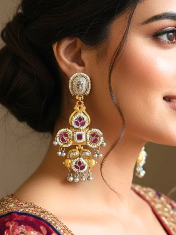 A close-up of a person wearing Zaynika Pink Earrings—large, ornate brass earrings with intricate detailing, white and Pink stones, and hanging silver beads. The person has long dark hair and is in a Pink patterned top.  Long Oxidized earrings, german silver earrings, navratri earrings.  Cross shaped earrings. 