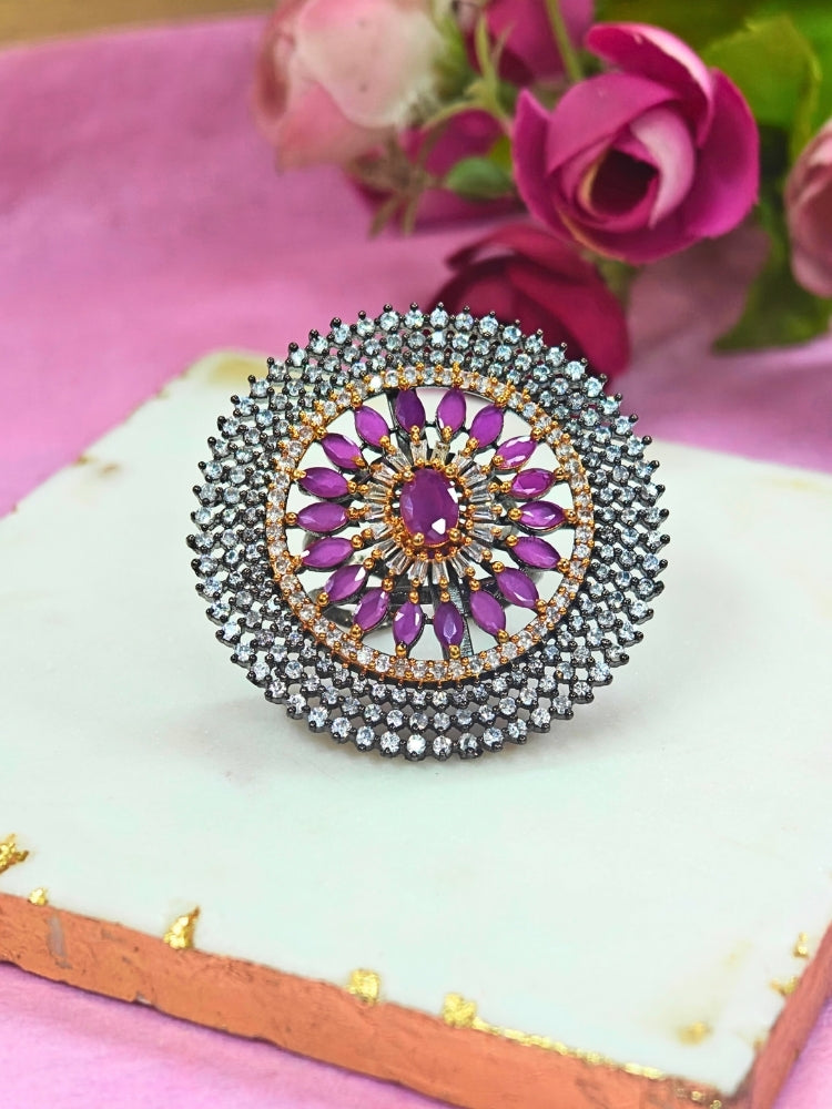 The Zenith Ring is a large, circular adjustable ring with a central purple gemstone, encircled by American diamonds and intricate smaller stones, displayed on white marble with pink and gold edges and flowers in the background.  Perfect American Diamond cocktail ring. 