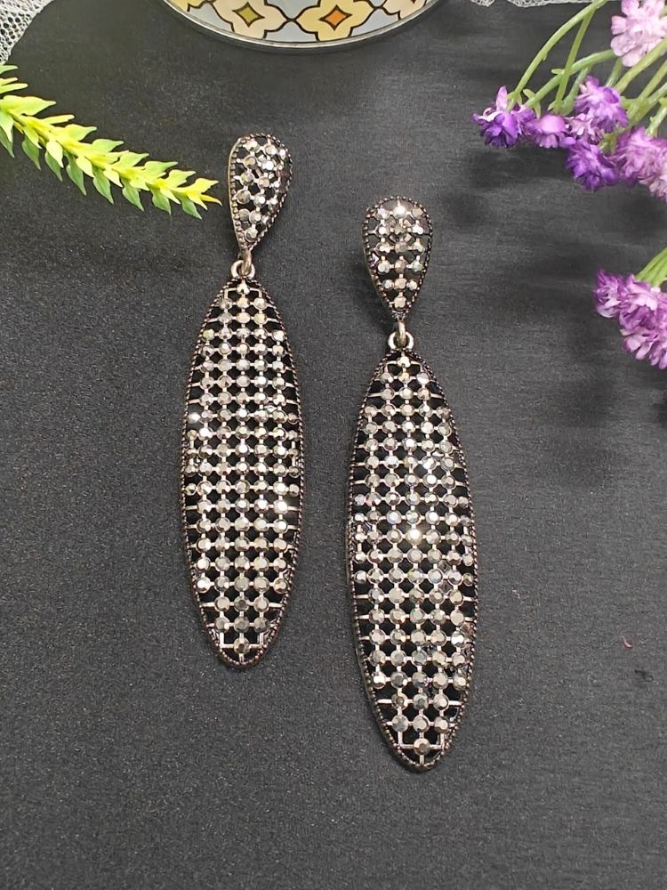 The Zeva Earrings are long, oval-shaped silver earrings with a teardrop top and a grid of black and crystal stones on a dark base, shown on black with purple flowers. Perfect for complementing your wedding ceremony dress.