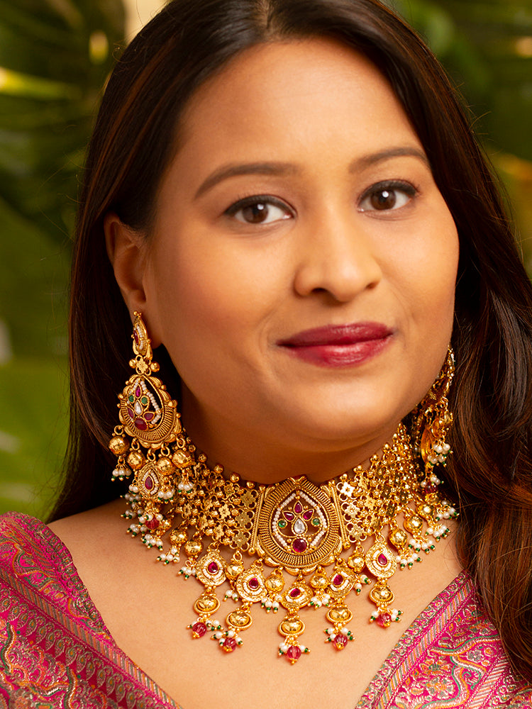 A woman in a pink and gold outfit smiles softly, wearing the Zoharika Choker Necklace Set—an elegant gold necklace set in jali work, with matching earrings adorned with red and white colored stones, ghungroo stones and more. Perfect for wedding outfits and the festive season. Blurred green foliage forms the background.