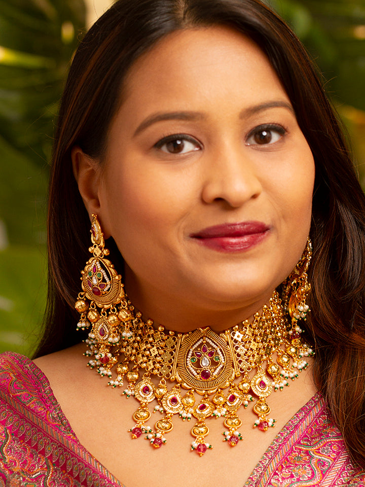 A woman in a pink and gold outfit smiles softly, wearing the Zoharika Choker Necklace Set—an elegant gold necklace set in jali work, with matching earrings adorned with red and white colored stones, ghungroo stones and more. Perfect for wedding outfits and the festive season. Blurred green foliage forms the background.